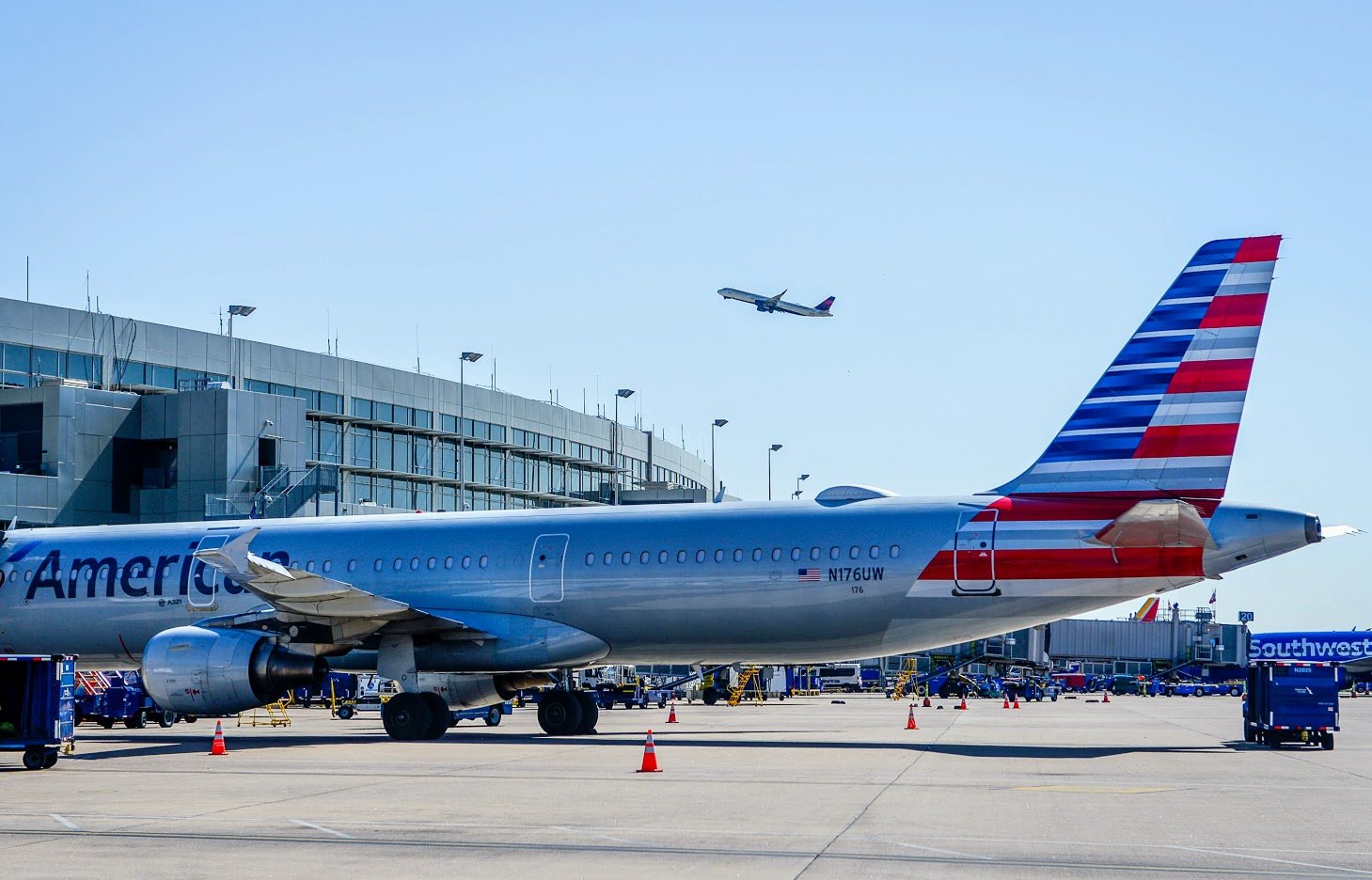 An American Airlines plane waits at the terminal gate. The plane is silver with an American flag painted on the tail. Another plane is in the sky above, having just taken off from the airfield.