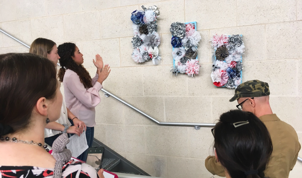 Students admire three floral looking three-dimensional art pieces hanging on a wall in a stairway.