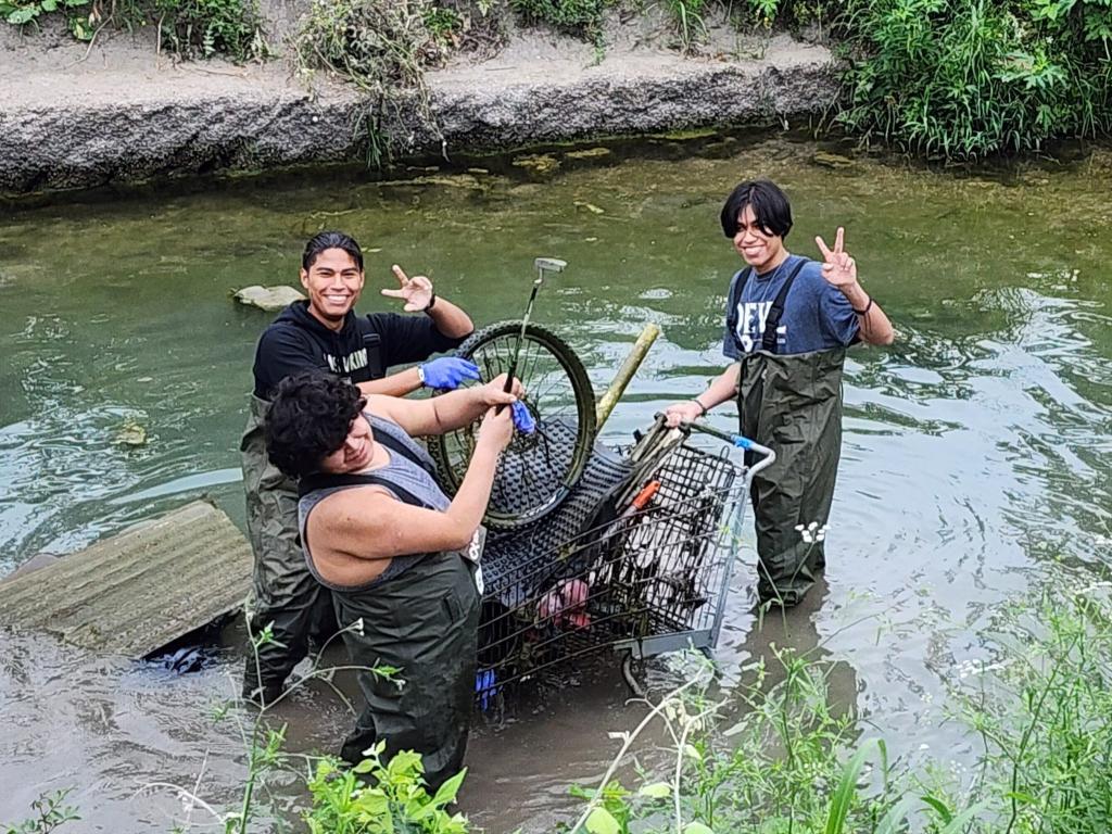 Three teenage volunteers remove a bicycle tire, shopping cart, and other trash from a creek