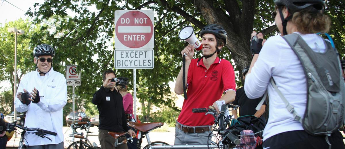 Cyclist with megaphone behind "Do not enter except bicyclists" sign. Council Member Chris Riley claps in the background.