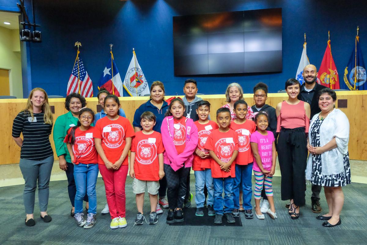 Kids with teachers stand in front of the City council dias. Most of the kids are wearing matching red shirts.