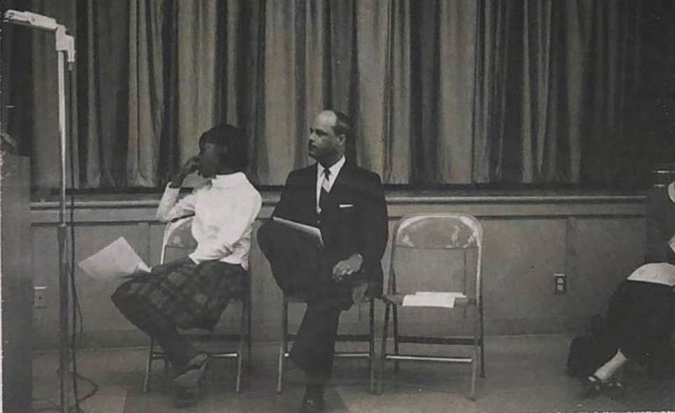 Picture of Dr. King seated on a folding chair beside a woman with a curtain behind 