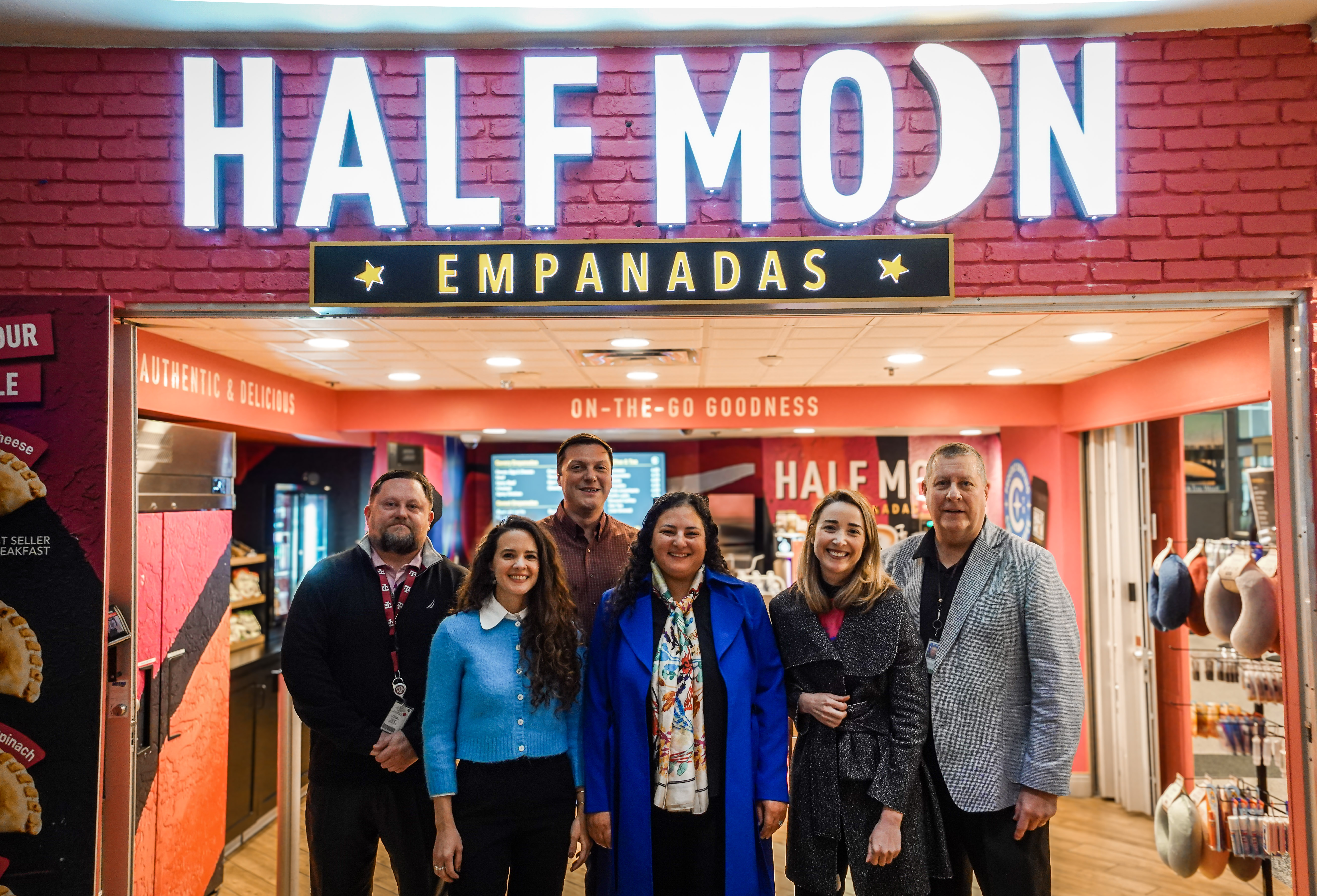 A group of seven people stands in front of the Half Moon Empanadas storefront, smiling for a photo. The illuminated sign above them reads "HALF MOON EMPANADAS," and the interior is decorated in red with signage promoting "Authentic & Delicious" and "On-The-Go Goodness." The individuals are dressed in business and business-casual attire, standing close together, celebrating the opening of the new food outlet.