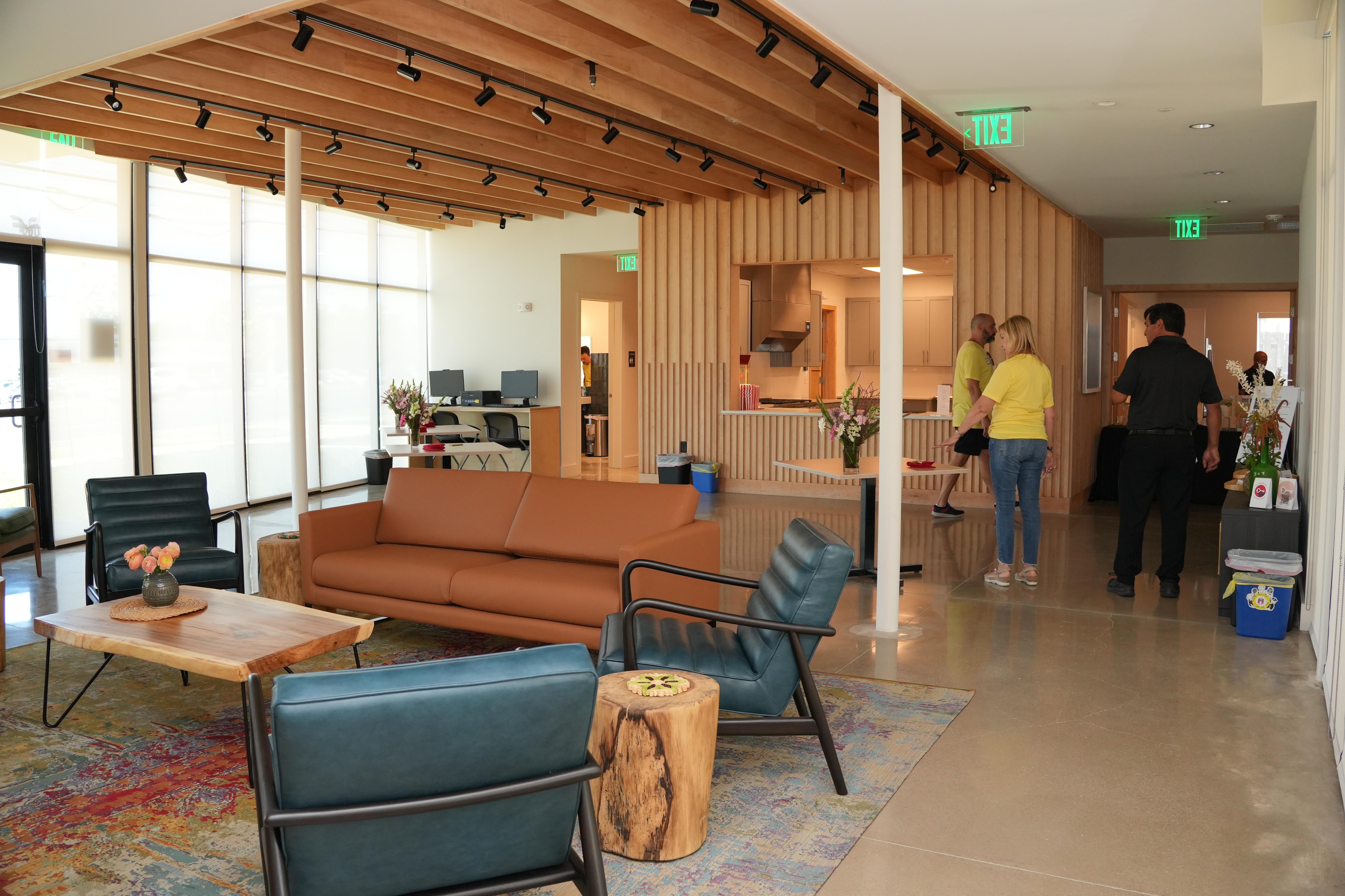 Image shows a modern interior common area with concrete floors, floor-to-celing windows, an orange leather couch, three blue leather side chairs, a wooden coffee table and two wooden end tables. 