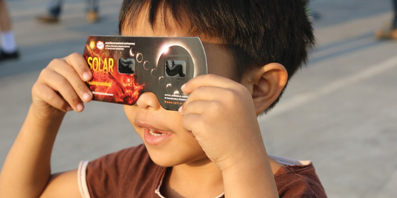 child holds solar eclipse glasses up to their eyes