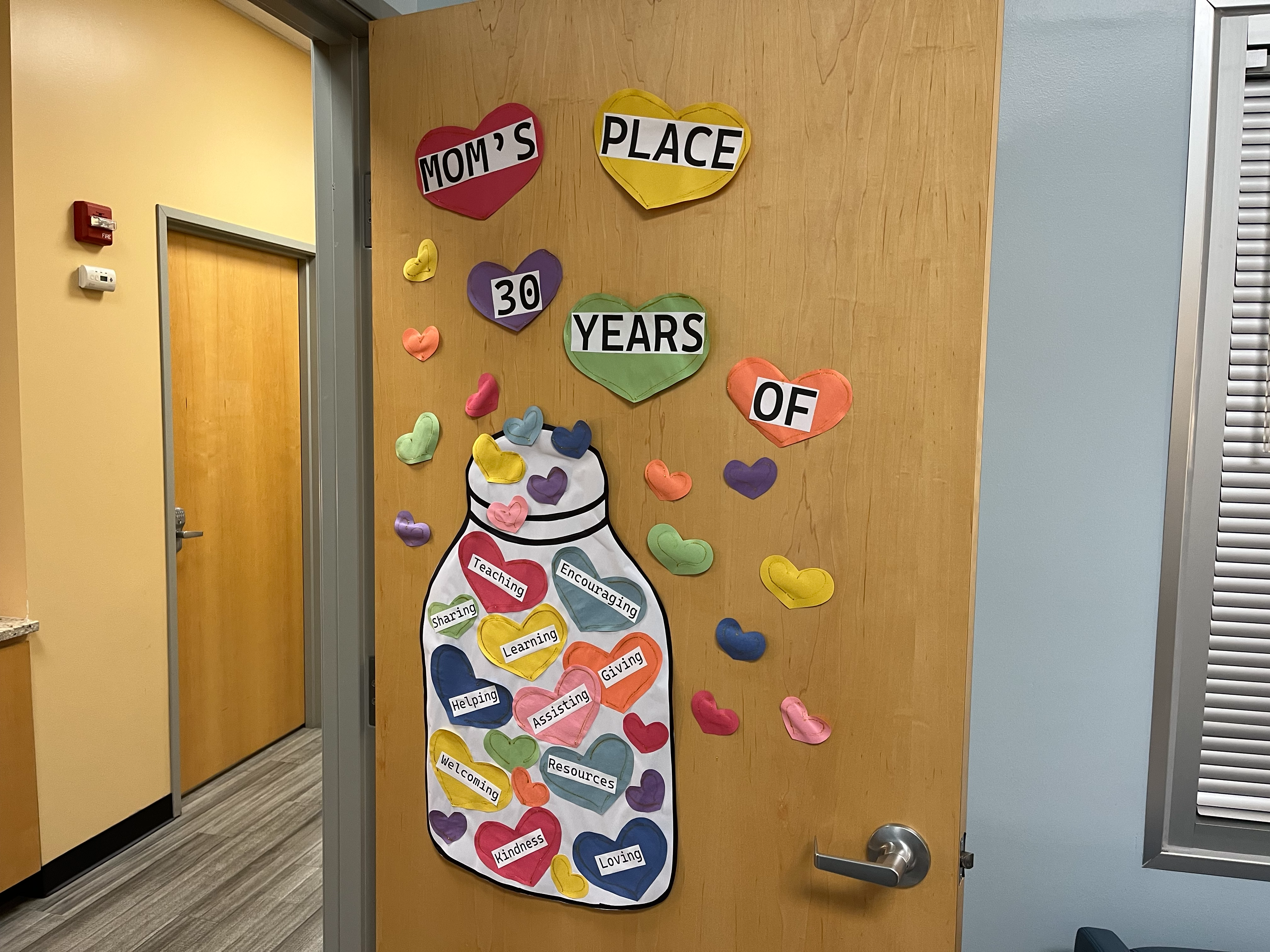 A door with art that reads Moms Place 30 Years of Teaching Encouraging Sharing Learning Giving Helping Assisting Resources Welcoming Kindness Loving