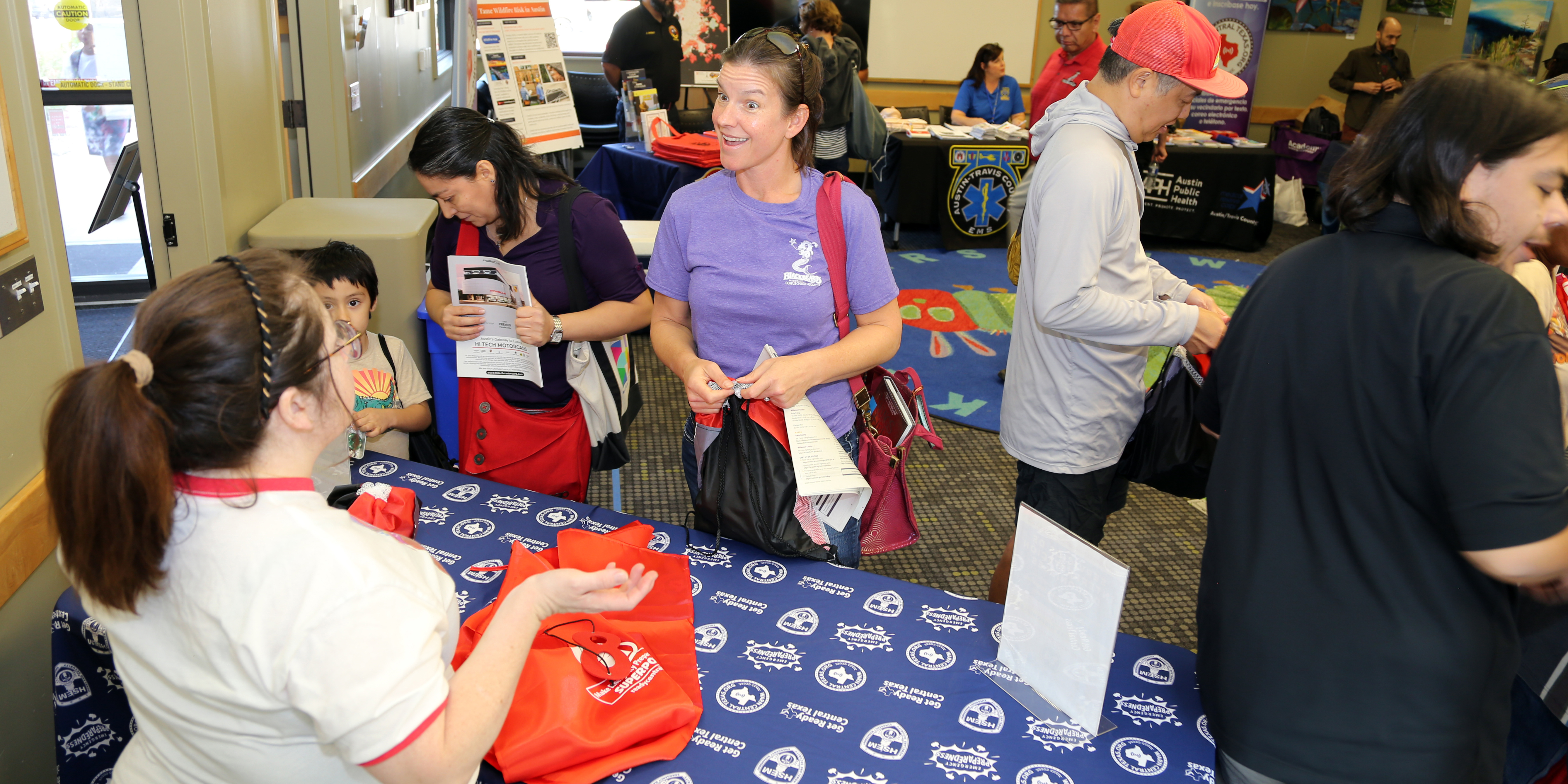 Caucasian woman smiling and speaking to another woman at a community outreach event with other people meeting and speaking to each other at tables.