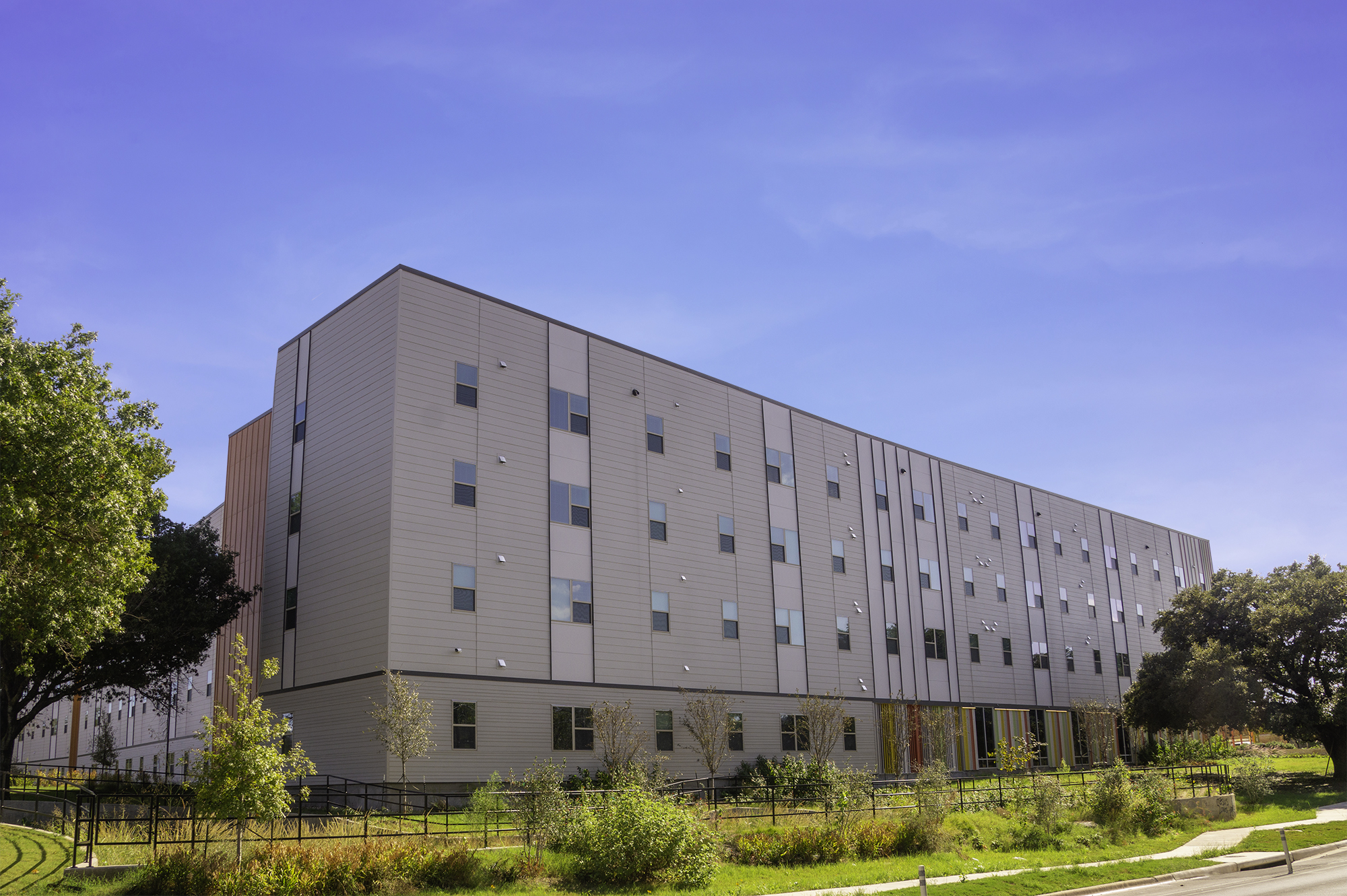 Photo shows an exterior view of the Parker Lane Apartment complex, including a four-story building that appears to be made of concrete masonry unit blocks. Also shown are trees and shrubbery and a black metal, handrail-lined walkway. 