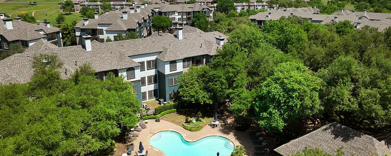 An aerial view of Twelve100, a residential complex, surrounded by lush green trees. The buildings have gray roofs and light-colored walls. A central courtyard features a large swimming pool with patio seating, creating a serene and inviting atmosphere.