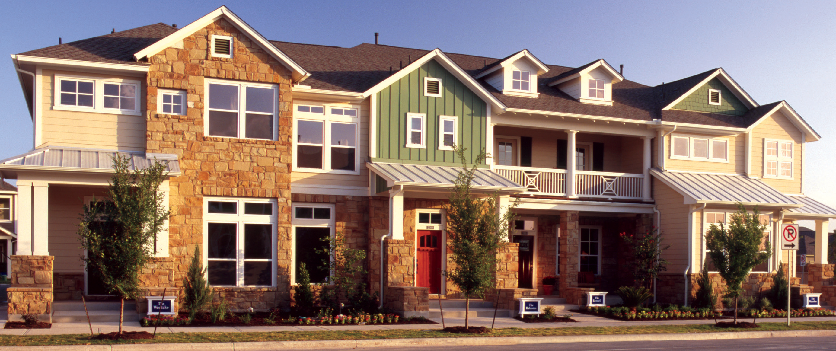 In Austin's Mueller neighborhood: A row of modern two-story townhomes with a mix of tan stone and light siding. The homes have large windows, red doors, and small landscaped gardens. Some parts of the buildings have green siding, and metal roofs cover the entryways. Young trees line the sidewalk, and signs in front of the homes display their names.