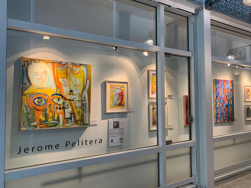 View through a glass gallery wall displaying an art exhibition. Colorful abstract portrait paintings hang in a row under spotlights, with a large expressive face painting on the left and smaller framed works to the right. The artist’s name, ‘Jerome Pelitera,’ appears in black lettering on the glass below the artworks.
