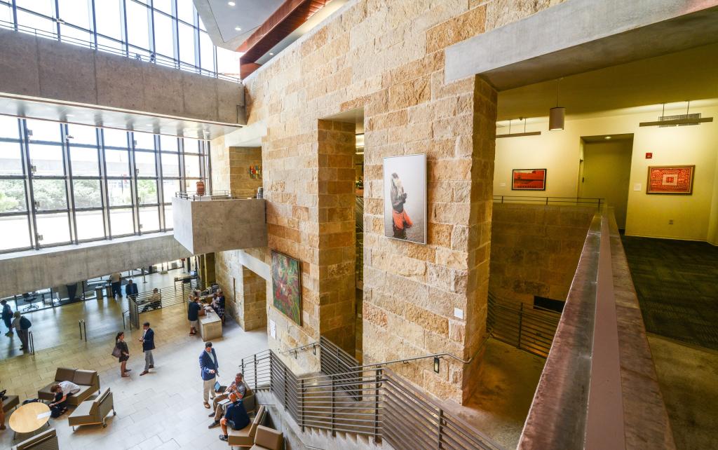 Interior of a modern multi-level atrium featuring stone walls, large glass windows, and concrete elements. Artwork is displayed on the walls, including a photo of a person in red pants and a colorful abstract painting. People are seated on the lower level, while the upper level has a hallway with more framed art. The space is bright, open, and designed for both function and aesthetics.