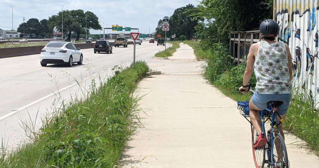 Person with helmet riding bicycle on aidewalk that is situated next to a frontage road