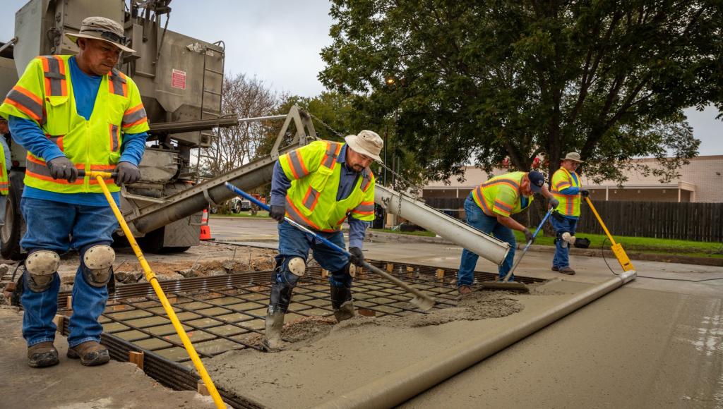 A crew of workers in yellow safety gear pour concrete on a street