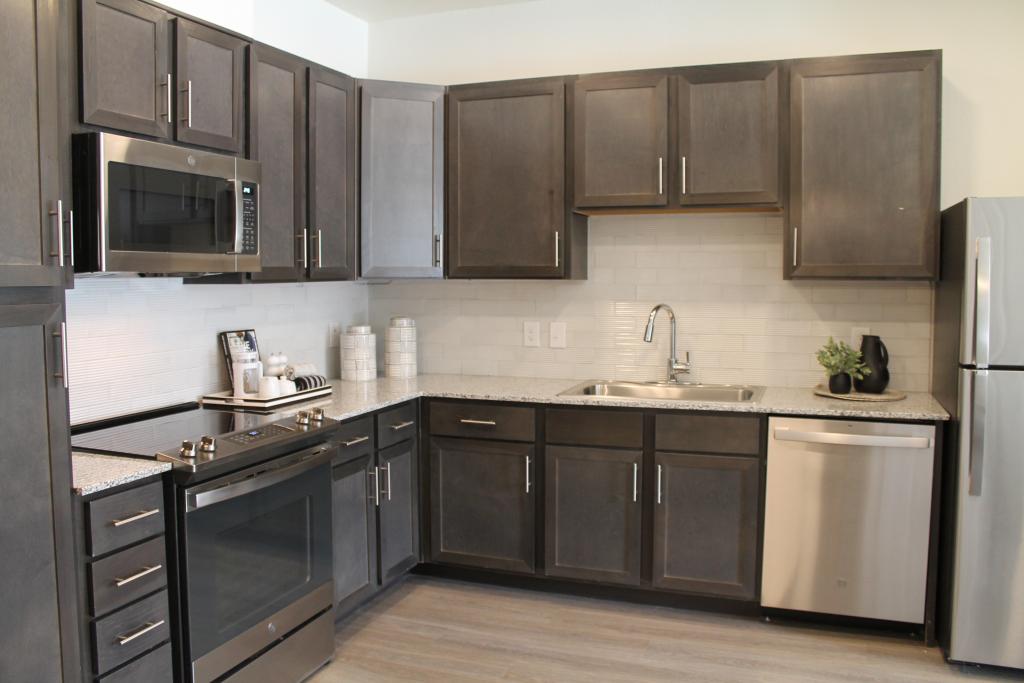 Photo shows the interior of a one-bedroom apartment at the Ekos City Heights residence facing the kitchen. The cabinets are a dark brown finish and there are stainless steel appliances. 