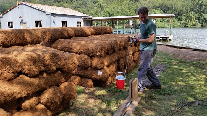 Coir logs, woven logs made of coconut husk fiber, are staged and prepped at Commons Ford Park for shoreline installation.
