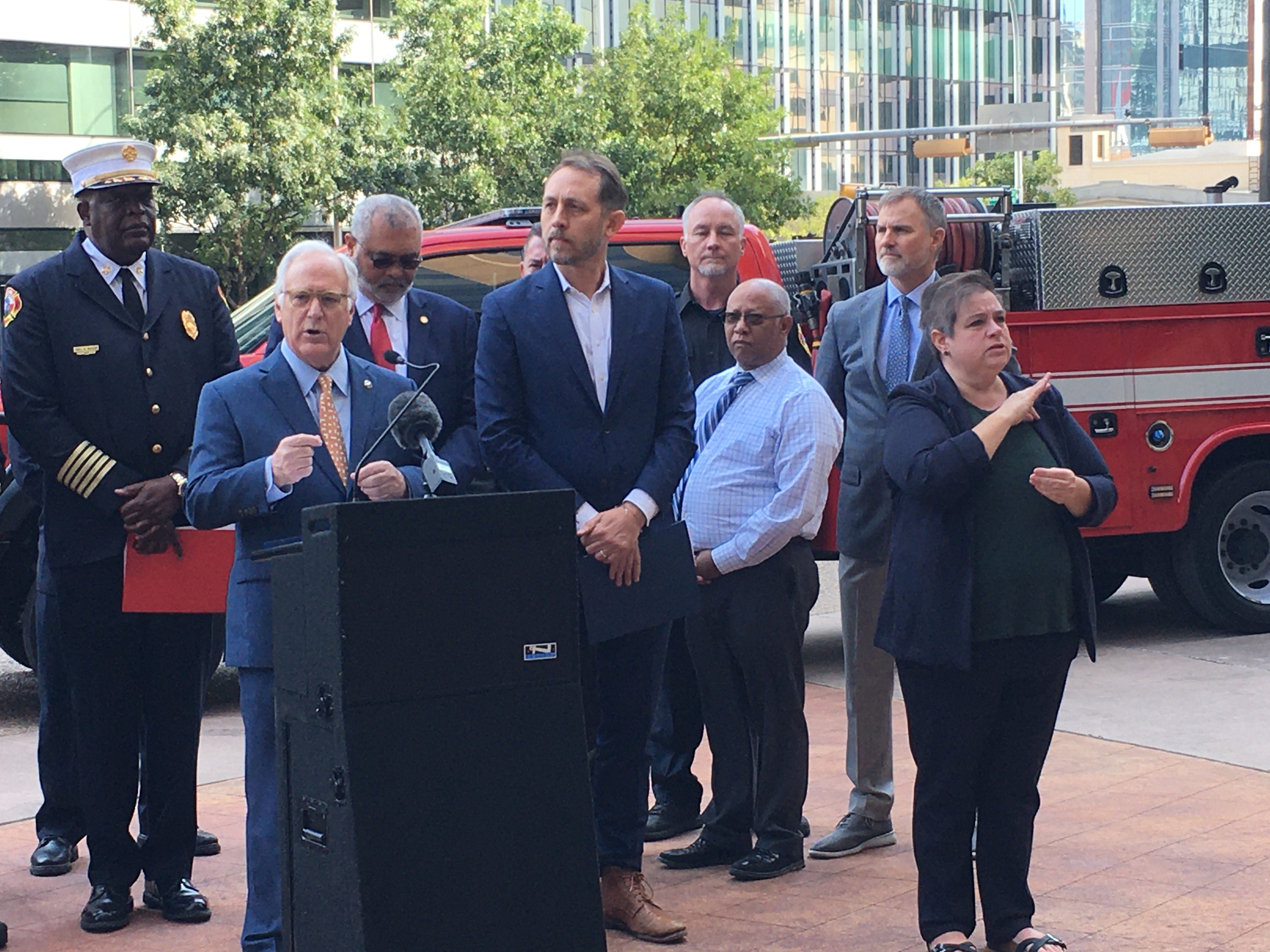 City of Austin Mayor Kirk Watson speaks at the podium during a press conference about wildfire danger. Other elected officials and fire chiefs stand behind him with 2 fire trucks in the background. A television camera is in the foreground.