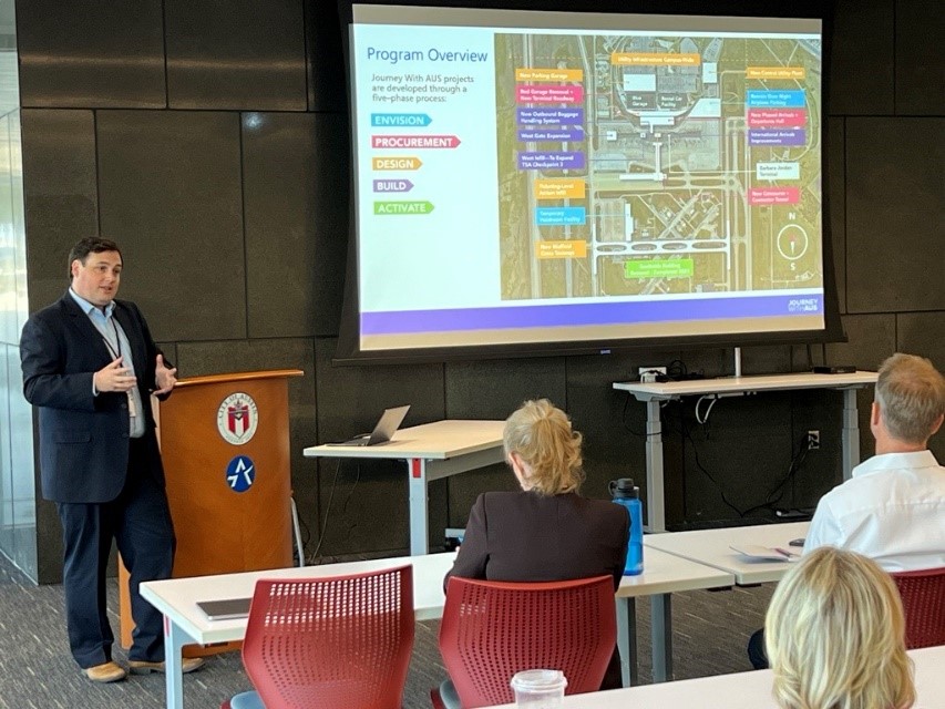 A person at the front presenting a slide show. People sitting at tables. 