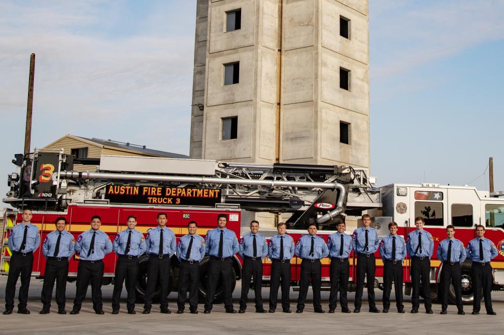 16 men wearing blue shirts, black ties and black pants with hands clasped behind backs, standing in front of Austin Fire Department engine