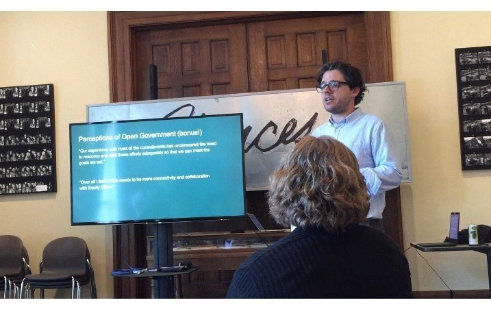 A photograph of a man next to a TV showing a slide deck on Perceptions of Open Government