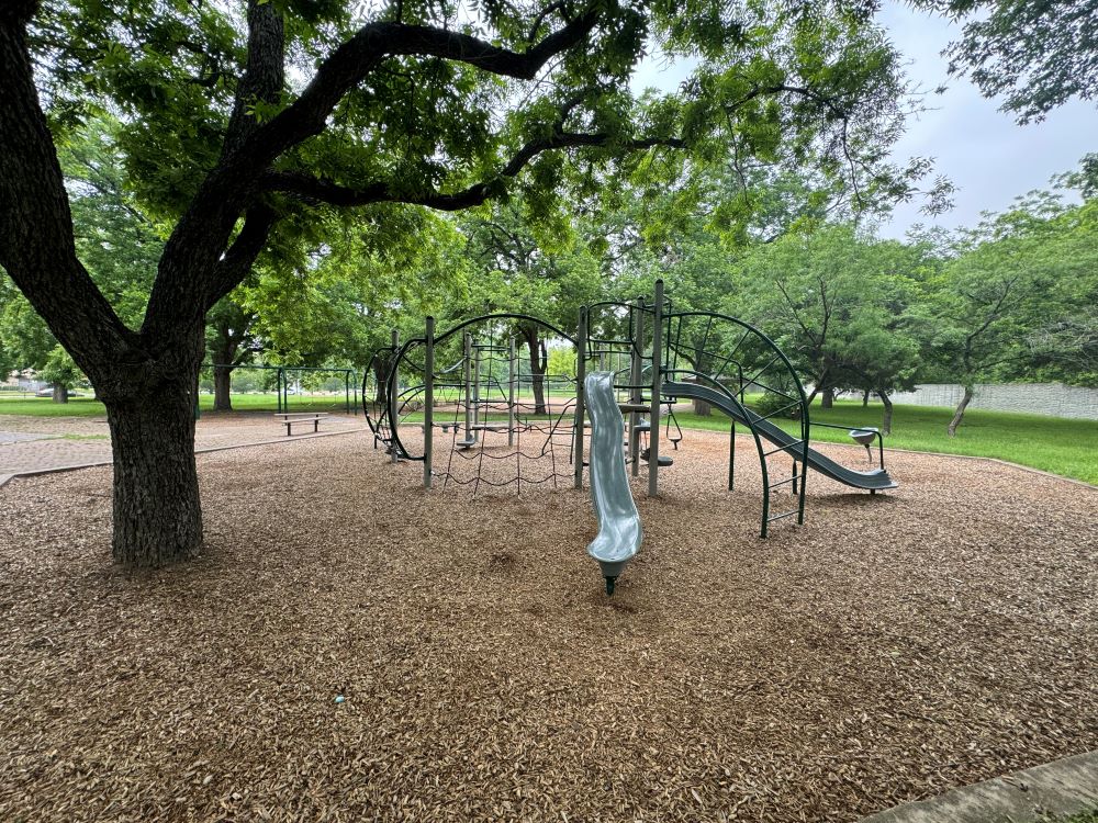 A metal playscape with slides and nets is shown in the foreground next to a tree.