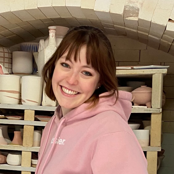  A close up portrait of a person smiling and making eye contact with the viewer. Behind them are ceramic artworks on a shelf.