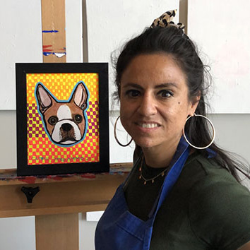 A photograph of a person close up they are smiling and making eye contact with the viewer next to them is a small easel with a painting of a dog