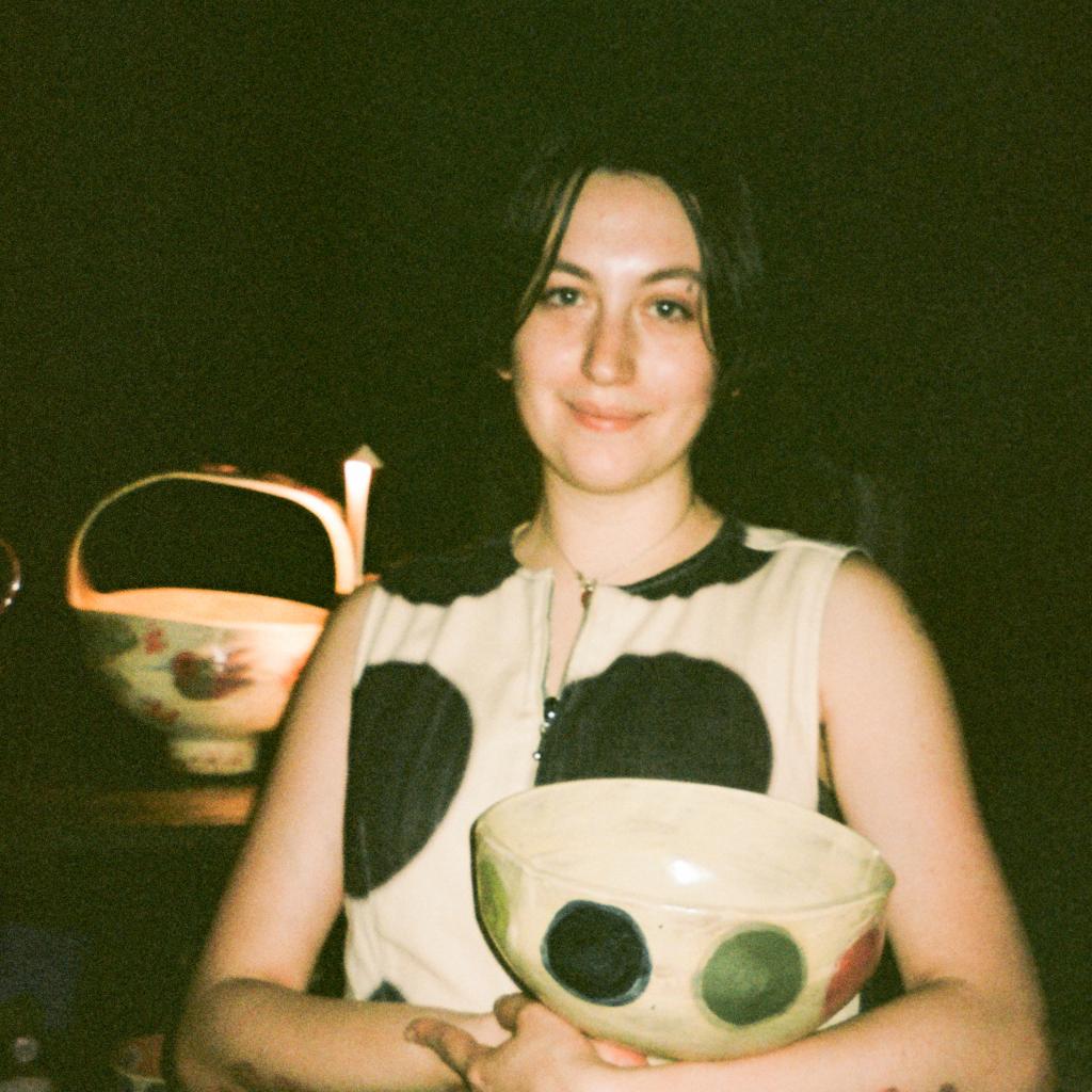 A photograph of artist Jamie Lerman holding a ceramic bowl, smiling, and making eye contact with the viewer