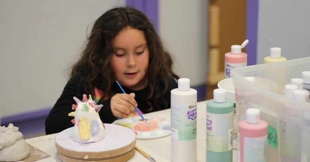 Youth student dips paintbrush into wet glaze in a paint palette to brush onto their multicolored ceramics project