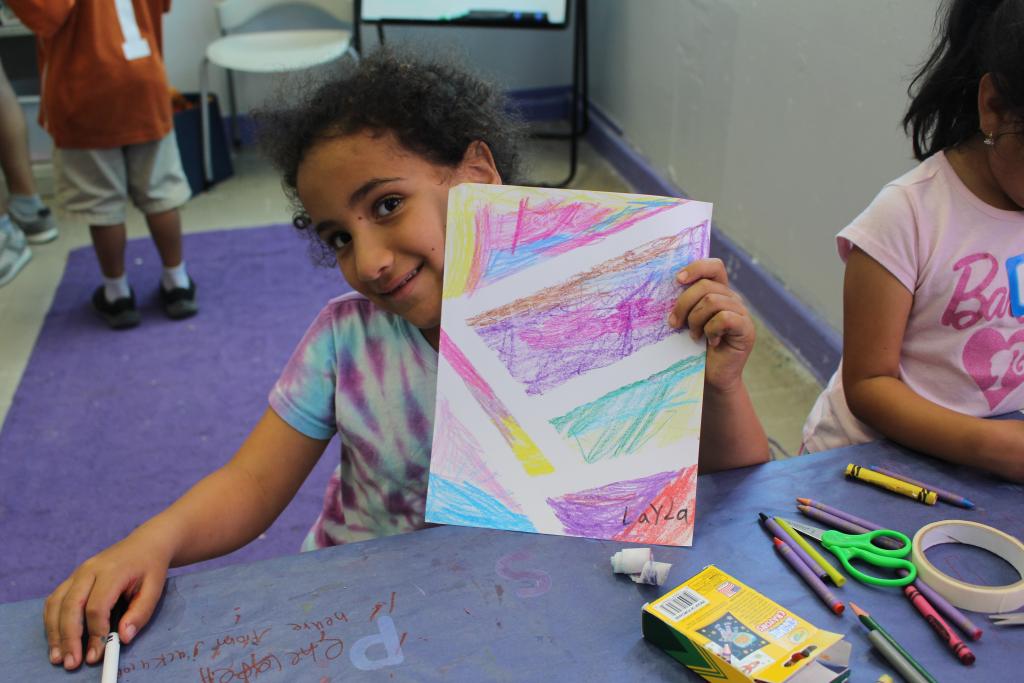 youth student seated at a table with various art supplies holds up colorful abstract artwork