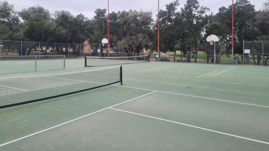 Image of shared-use courts at South Austin Neighborhood Park showing pickleball court and net, tennis court and net, basketball goals, trees in background