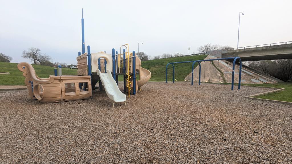 Boat themed playground with swings nearby and roadway behind the playground