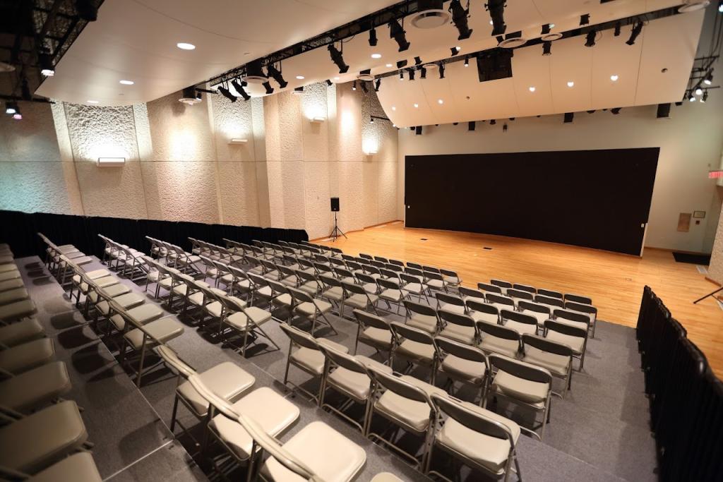 A spacious auditorium with a wooden stage and tiered seating filled with rows of gray folding chairs. The walls have a textured design, and the ceiling features multiple stage lights. A black backdrop is at the front of the stage, and a speaker on a stand is positioned to the side.