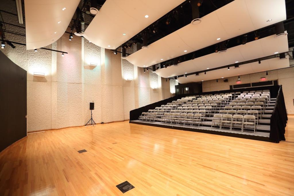 A wide view of an auditorium with a polished wooden floor and a large seating section arranged on a raised platform. The ceiling is equipped with stage lighting, and the walls have a textured finish. A black speaker stands near the stage, and multiple exit doors are visible in the background.