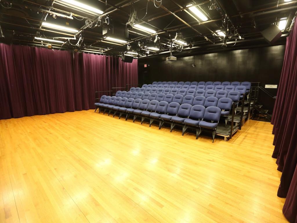 A black box theater with a wooden floor and tiered seating featuring rows of cushioned blue chairs. The space is surrounded by dark walls, and the ceiling is equipped with multiple stage lights. The back of the room has deep purple curtains, partially covering the walls.