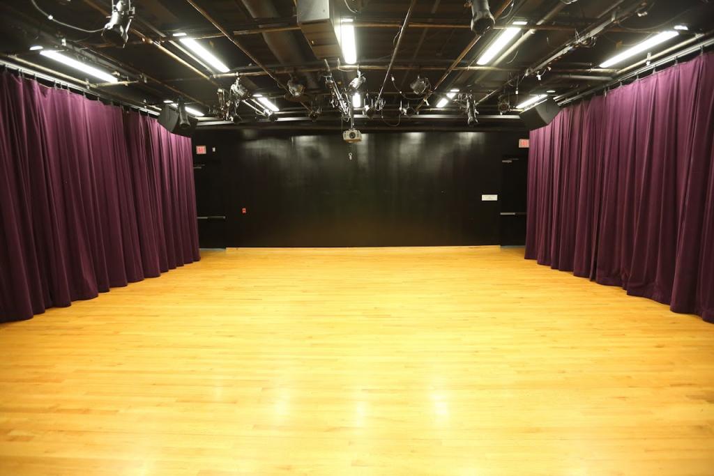 An empty black box theater with a polished wooden floor and dark walls. Deep purple curtains line the sides, and the ceiling is equipped with overhead stage lights and a projector. The room has a flexible setup, suitable for performances or events.