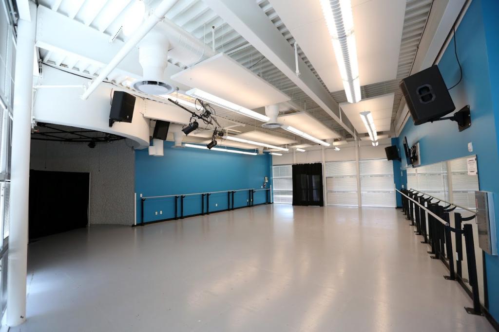 A spacious dance studio with a light gray floor, bright blue walls, and large windows with natural light. The studio features ballet barres along the walls, mounted speakers, and overhead lighting. A black curtain covers a portion of the space, and stage lights are installed on the ceiling.