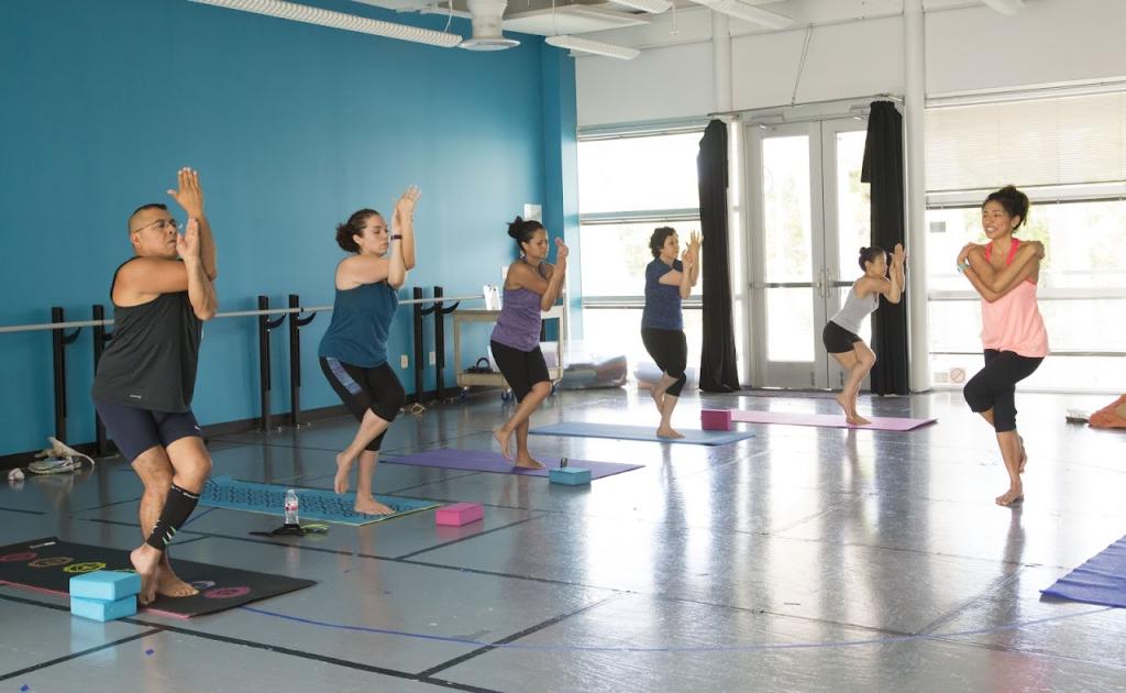 A group of five people participating in a yoga class inside a dance studio. They are standing on colorful yoga mats, following an instructor in a pink top, all performing the Eagle Pose. The studio has blue walls, ballet barres, and large windows with black curtains letting in natural light.