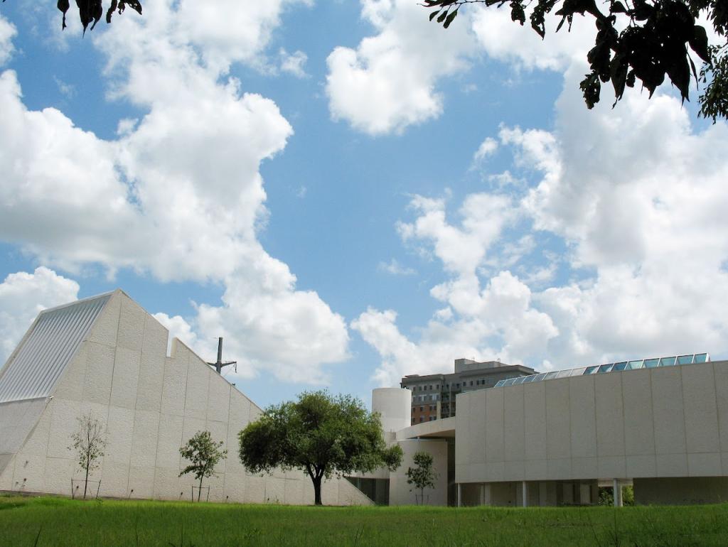  A modern white building with angular architecture surrounded by a green lawn and trees under a bright blue sky with fluffy clouds. A taller building is visible in the background.