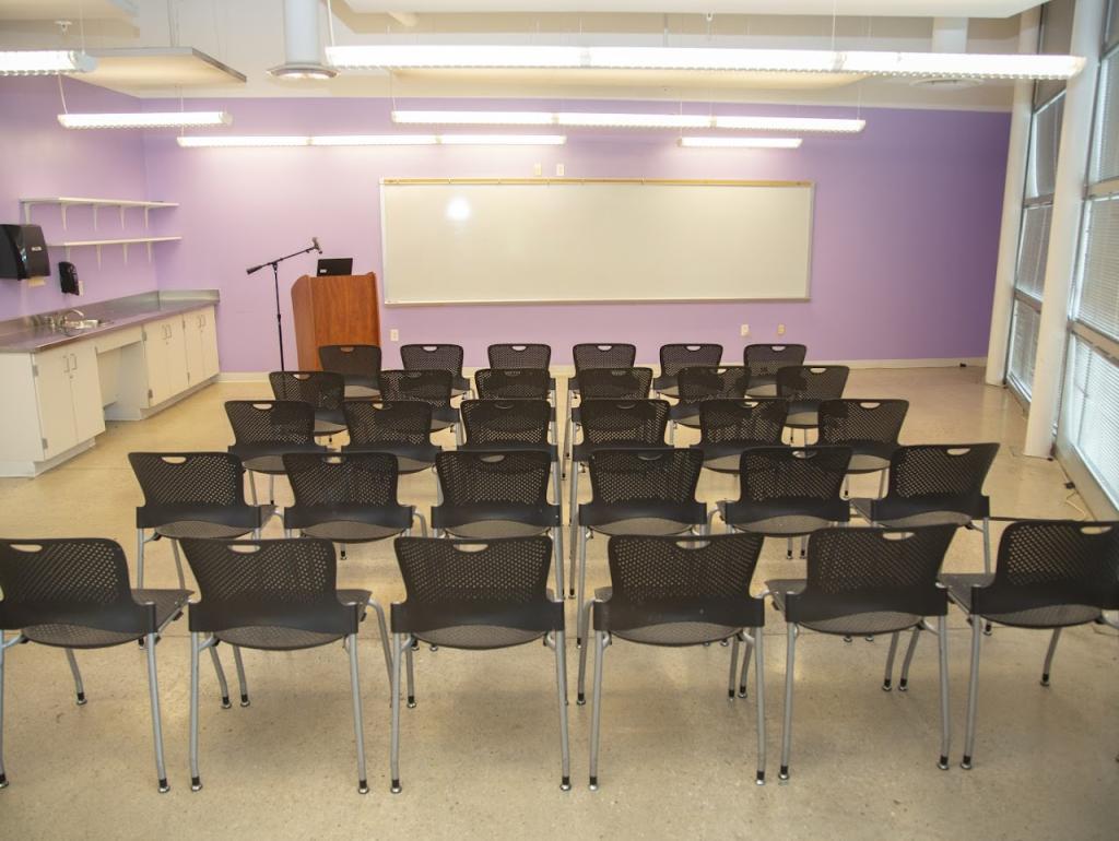 A classroom-style event space with purple walls, a whiteboard, and rows of black chairs facing a wooden podium with a microphone. The room features built-in cabinets, a sink, and large windows allowing natural light to enter.