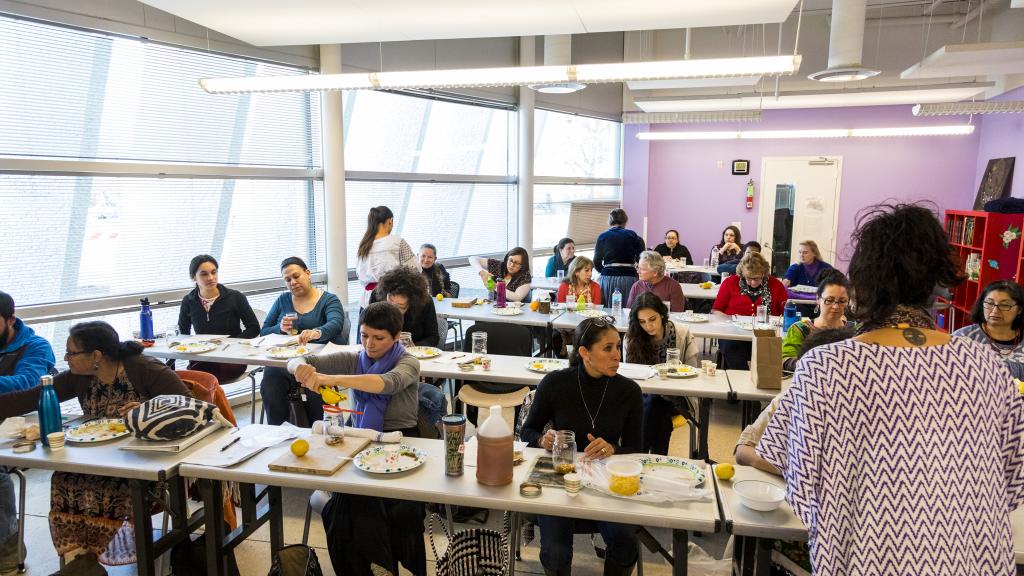 A group of people participating in a hands-on workshop inside a classroom. Participants are seated at tables covered with plates, cups, and jars as they engage in an interactive learning session led by an instructor at the front.