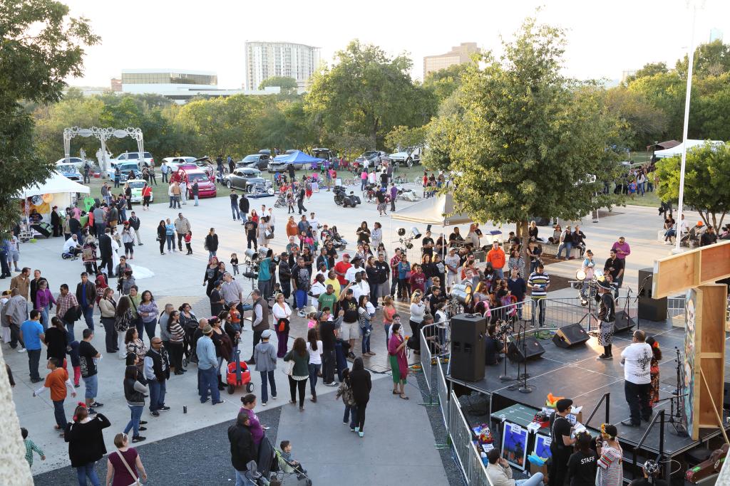 A lively outdoor festival with a diverse crowd gathered around a stage where a performance is taking place. People are walking around, socializing, and exploring booths and classic cars on display. The background features trees, tents, and high-rise buildings.