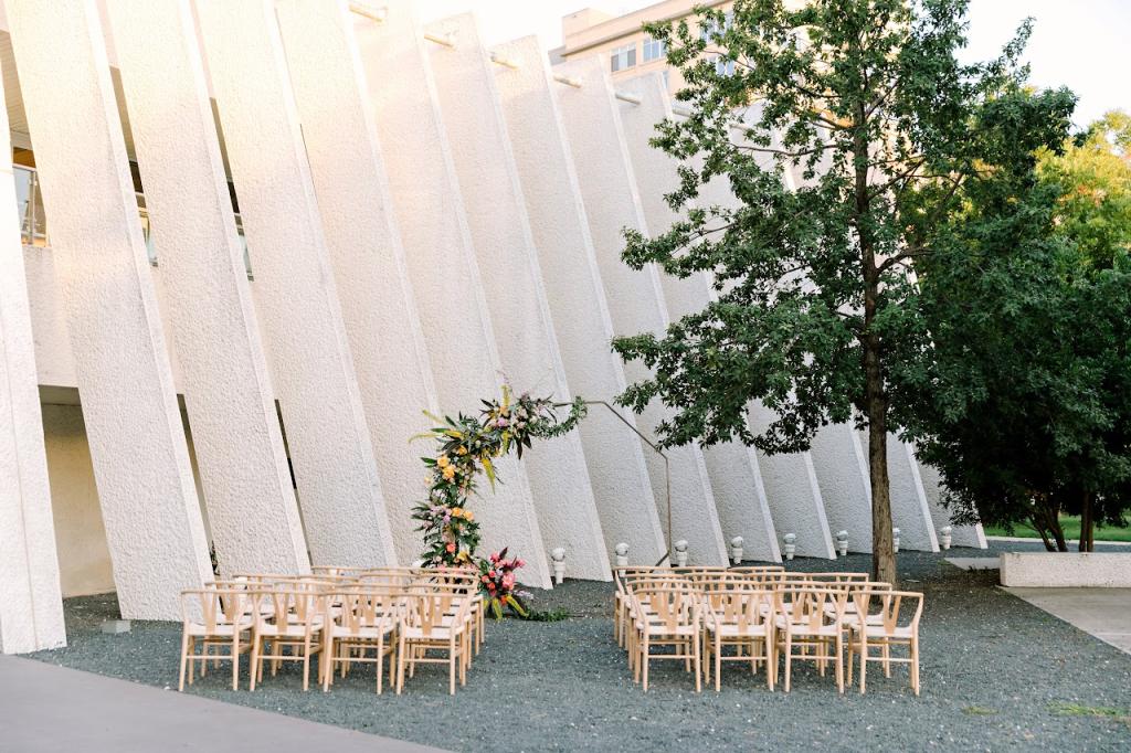 A beautifully arranged outdoor wedding ceremony setup with wooden chairs facing a floral arch. The backdrop features a modern white building with angled architectural panels, a tree, and soft natural lighting.