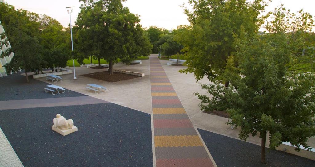 A paved outdoor plaza with a multicolored brick pathway lined by lush green trees. White picnic tables and benches are scattered throughout, and a white abstract sculpture is placed near the walkway.