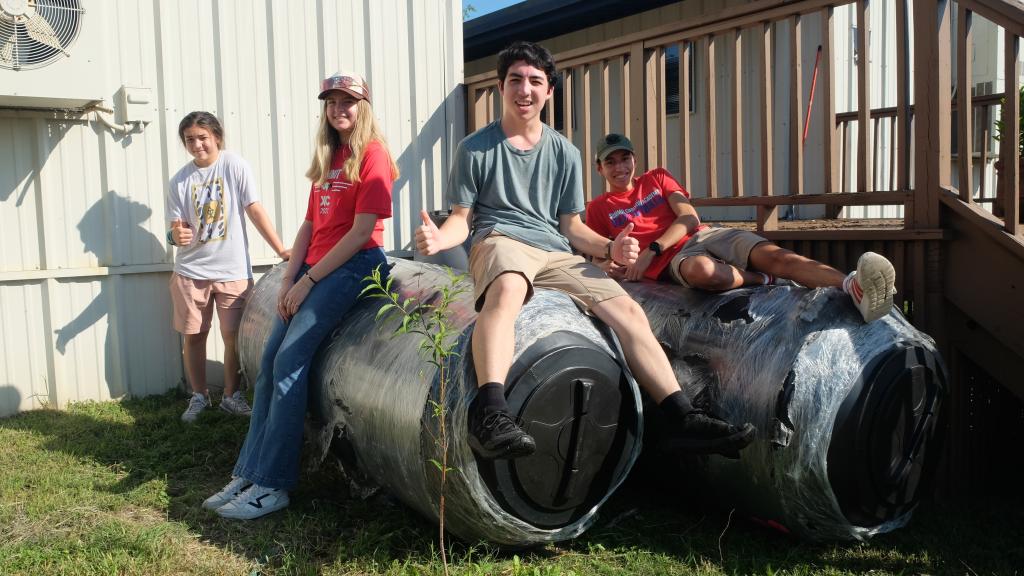 Four students sit on large rain barrels waiting to be installed.