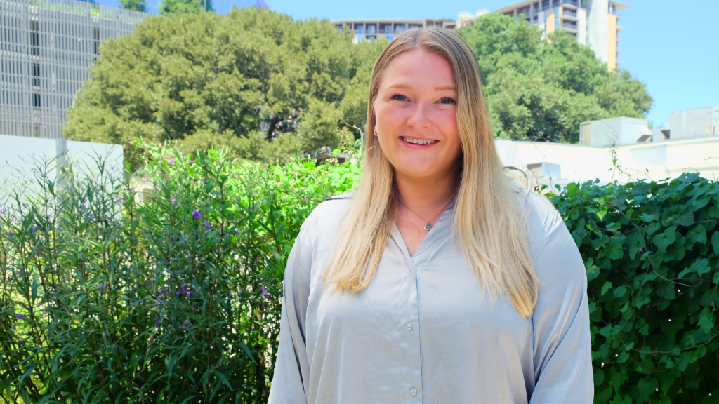 A photo of Molly Ellsworth smiling in front of local plants. Behind her are trees and buildings, one with a green roof.