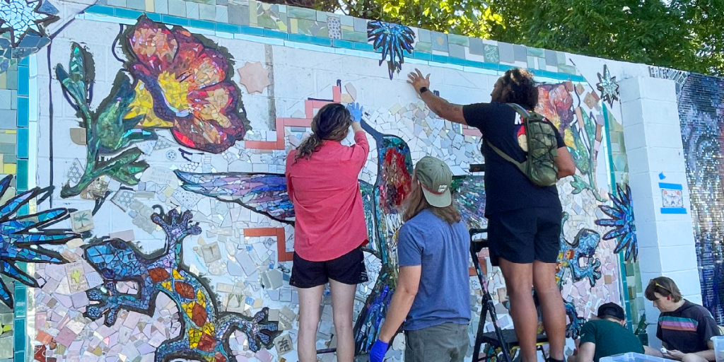 Five folks work on the Under the Sun mural. Some stand on ladders working on grouting mosaic pieces.