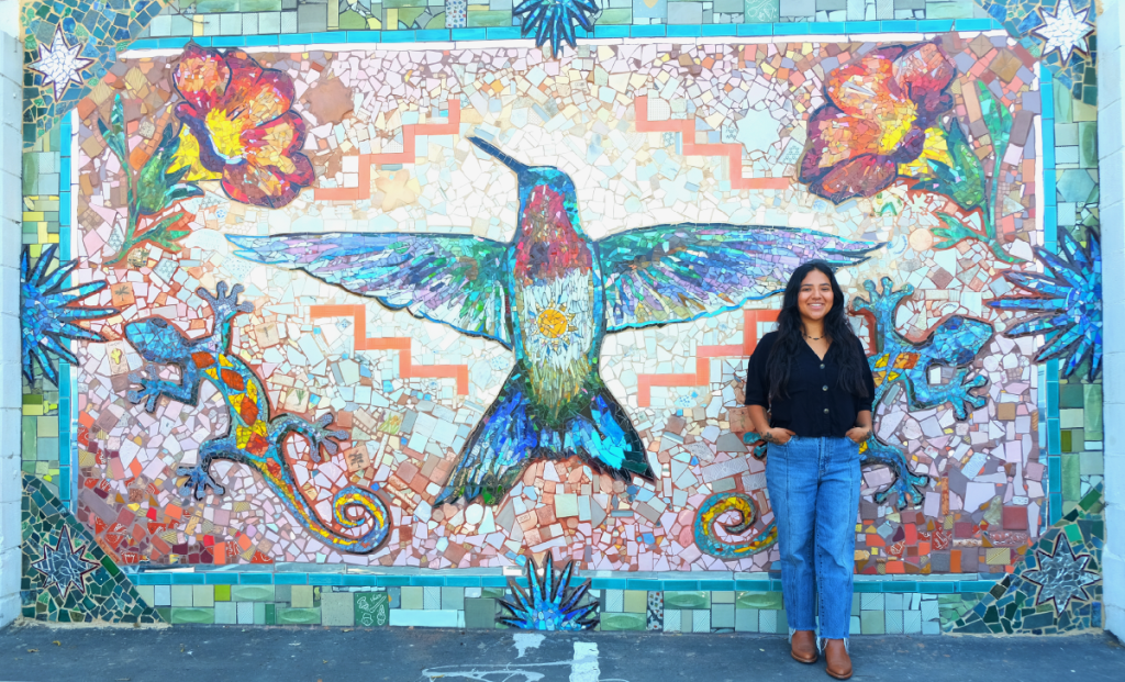 Carmen Rangel poses in front of large mural project featuring a hummingbird, flowers, and a gecko.