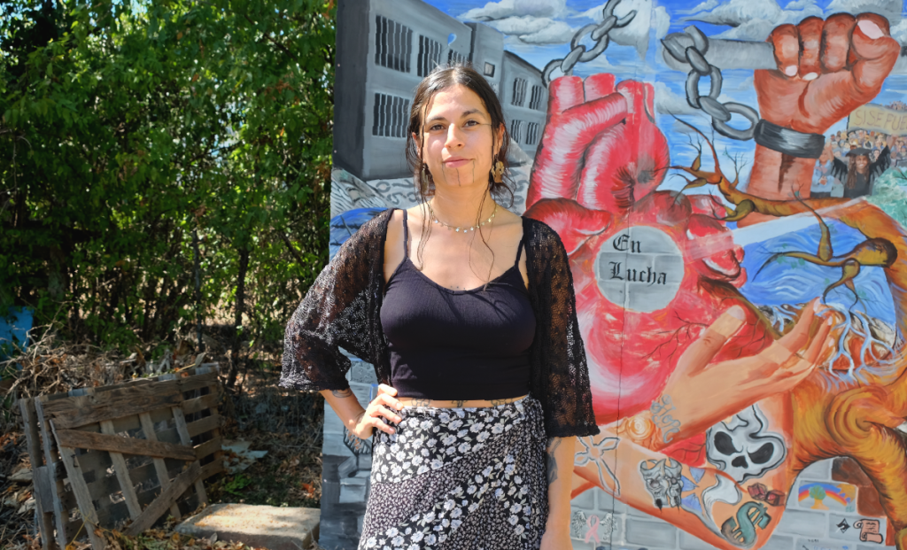 Chiara looks straight at the camera with one hand on her hip. Behind her is a mural depicting social justice themes.