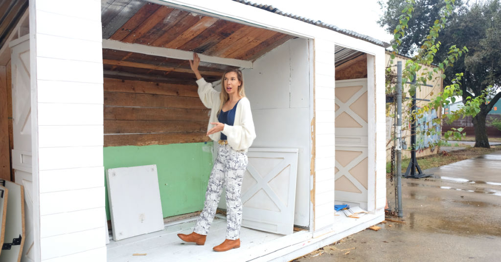 Kelley stands in a wooden framed structure outside with various tools and wooden pieces within it.